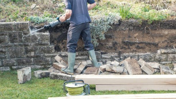 Man using a jackhammer to remove stone wall. He is wearing work boots and pants, working outdoors.