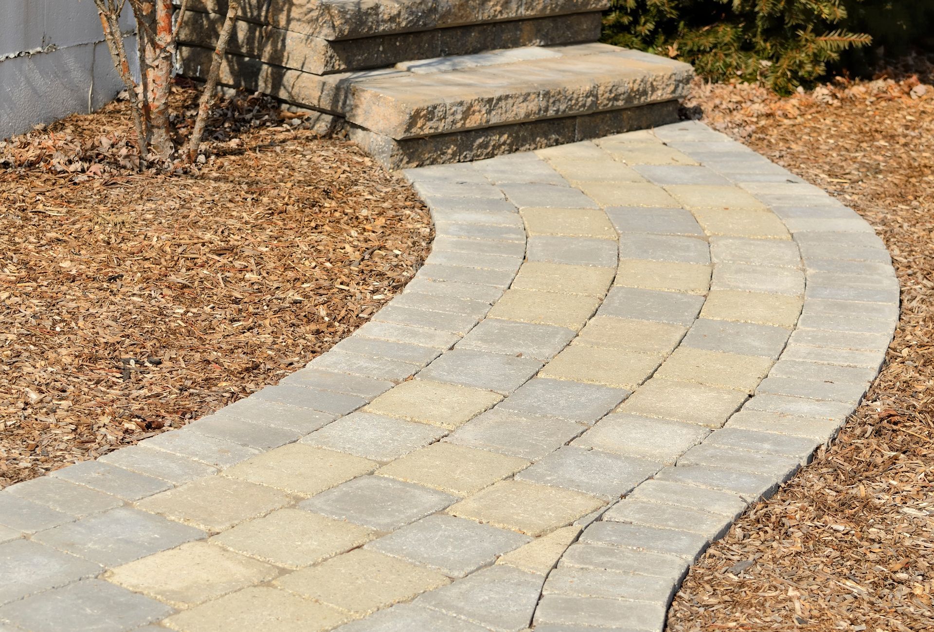 Stone path curves toward steps; tan and gray pavers, edged by brown mulch and grass.