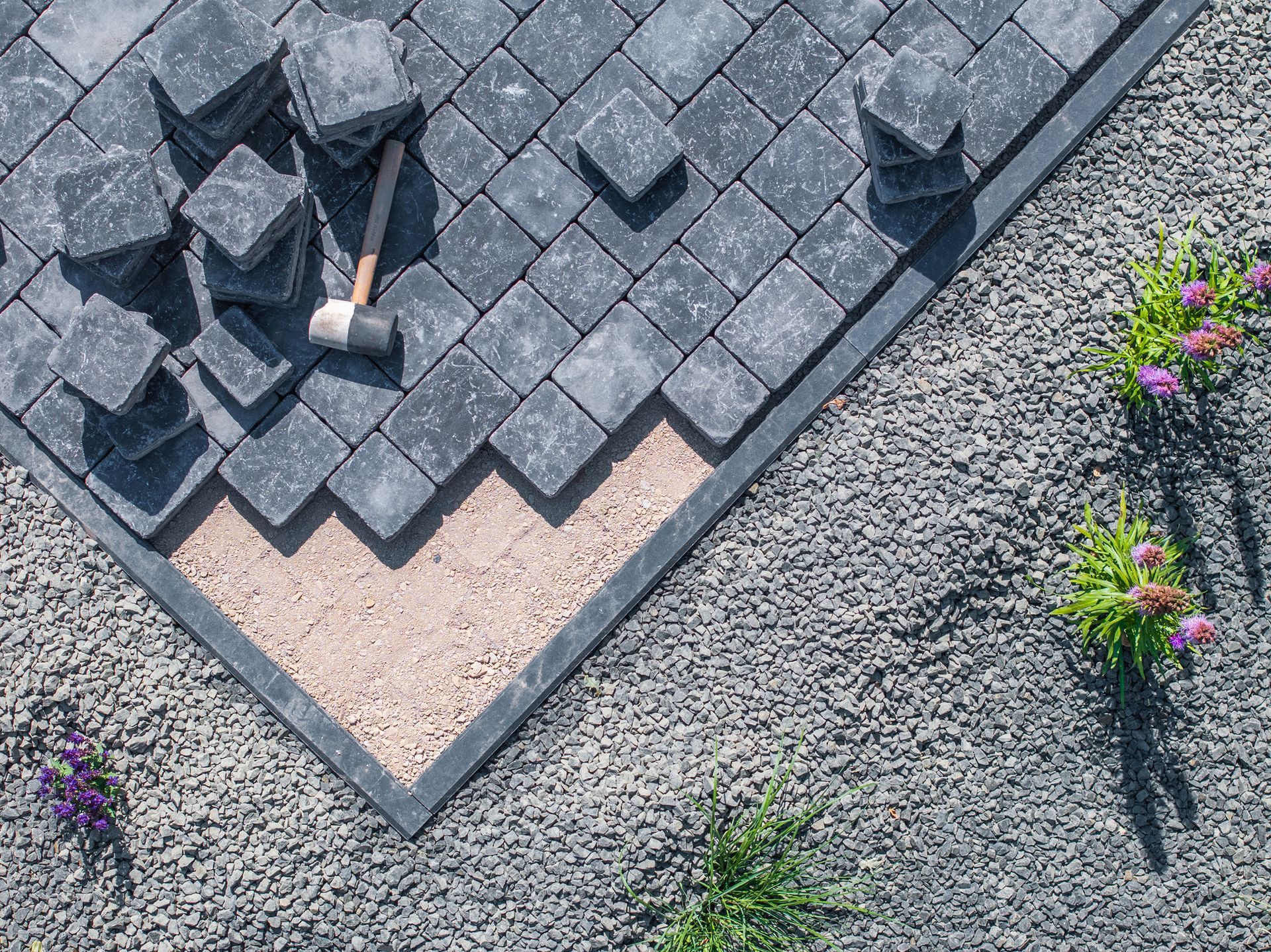 Installing pavers: Gray square stones being laid on sand base with a mallet, near gravel and plants.