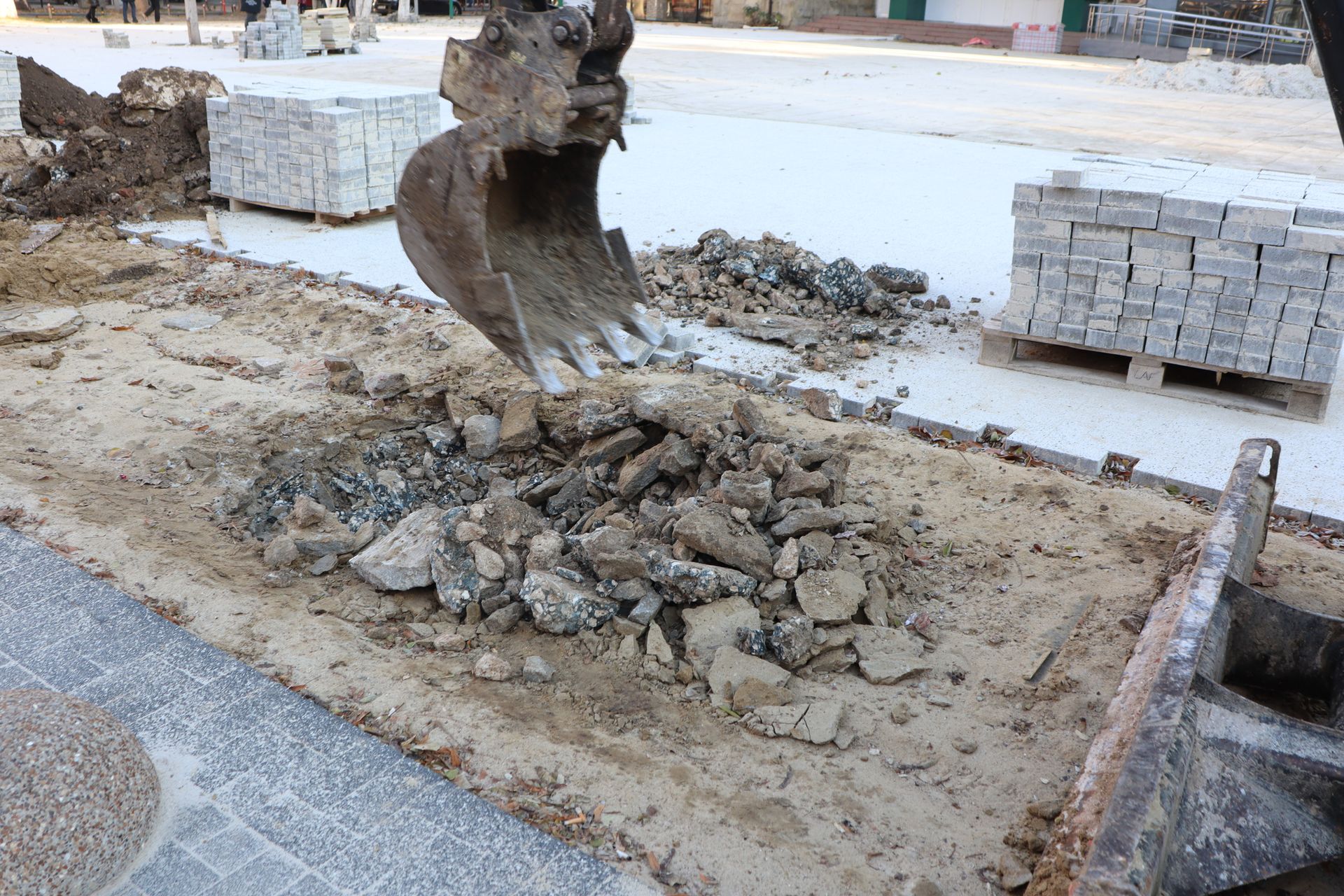 An excavator scoops rubble from a sidewalk, pallets of pavers in the background. An excavator scoops rubble from a sidewalk, pallets of pavers in the background.