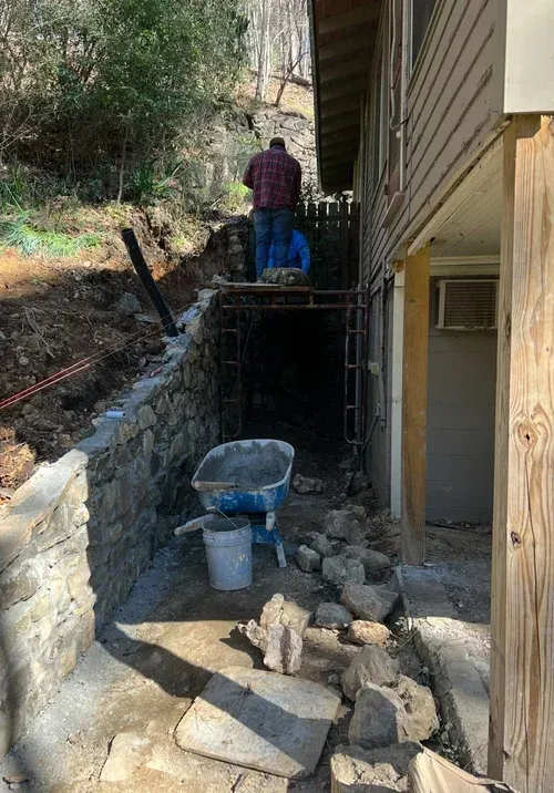 Construction site: Man working on a wall with a wheelbarrow, rocks, and scaffolding next to a building.