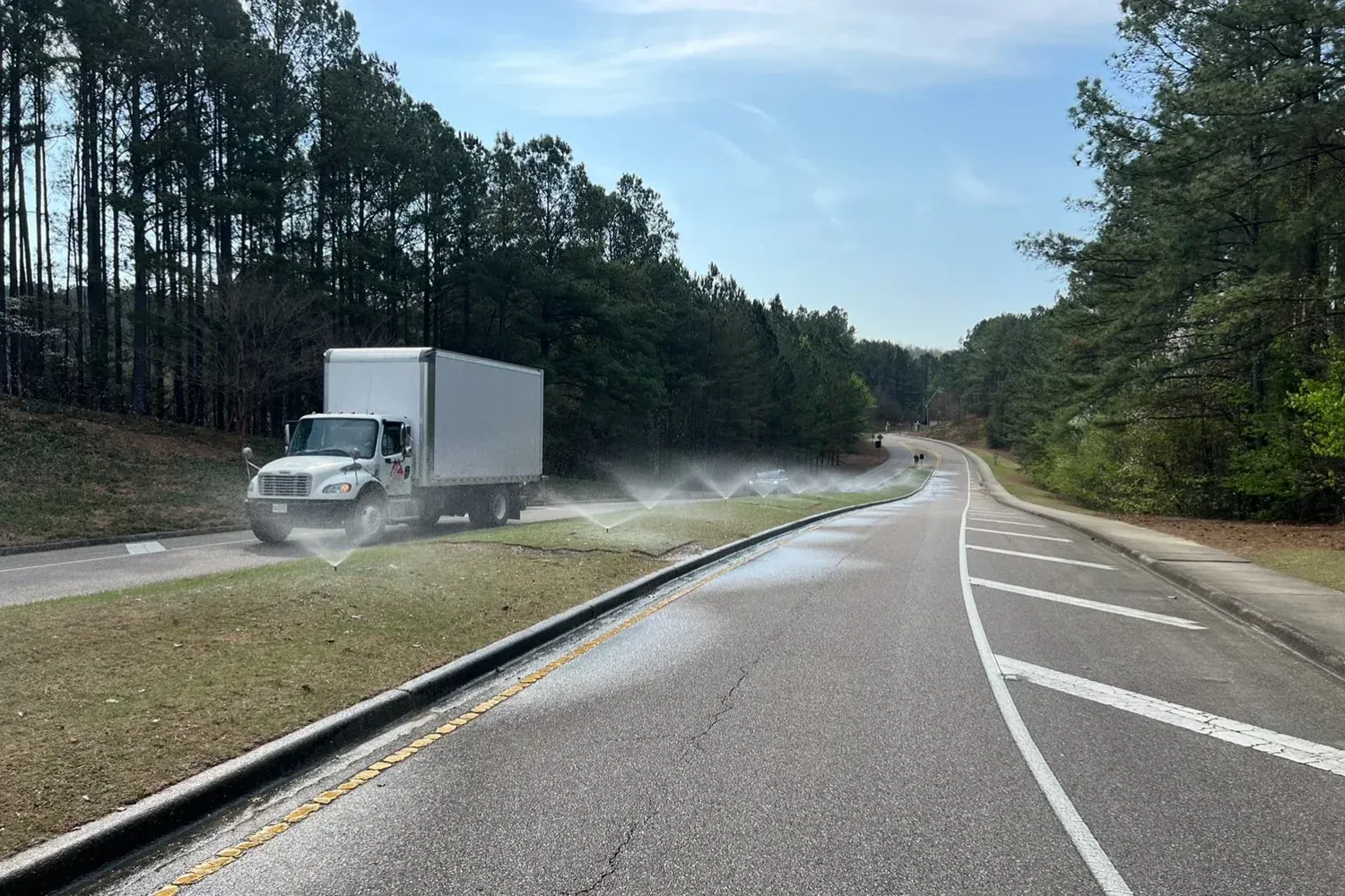 White truck spraying water on grass next to a road, near a forest.