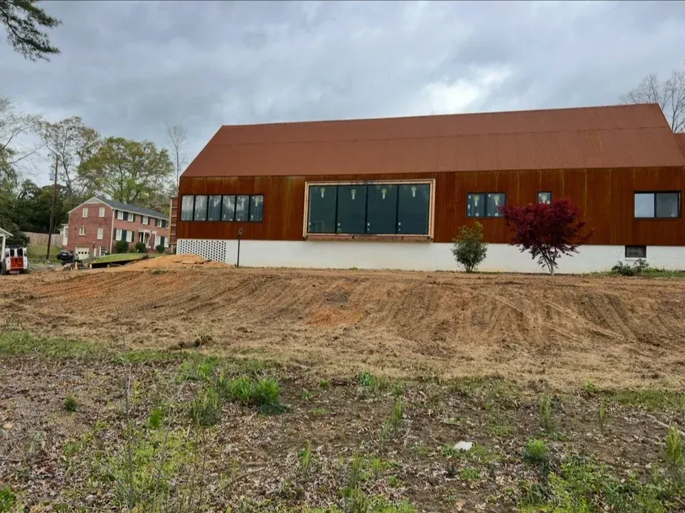 Modern brown building with large windows, under construction on a dirt lot, with an older brick building in the background.