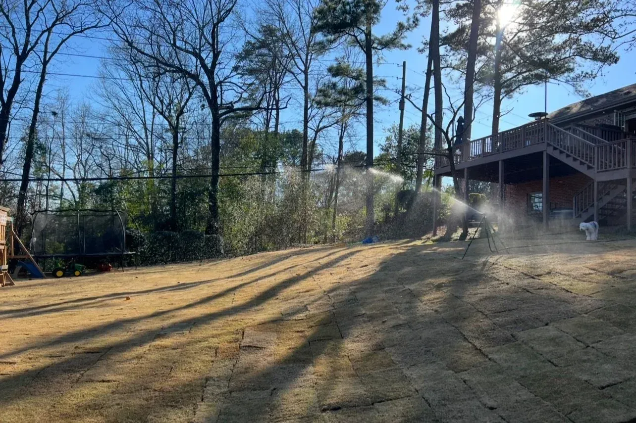 Sprinklers watering a lawn, near a house with a deck, on a sunny day.
