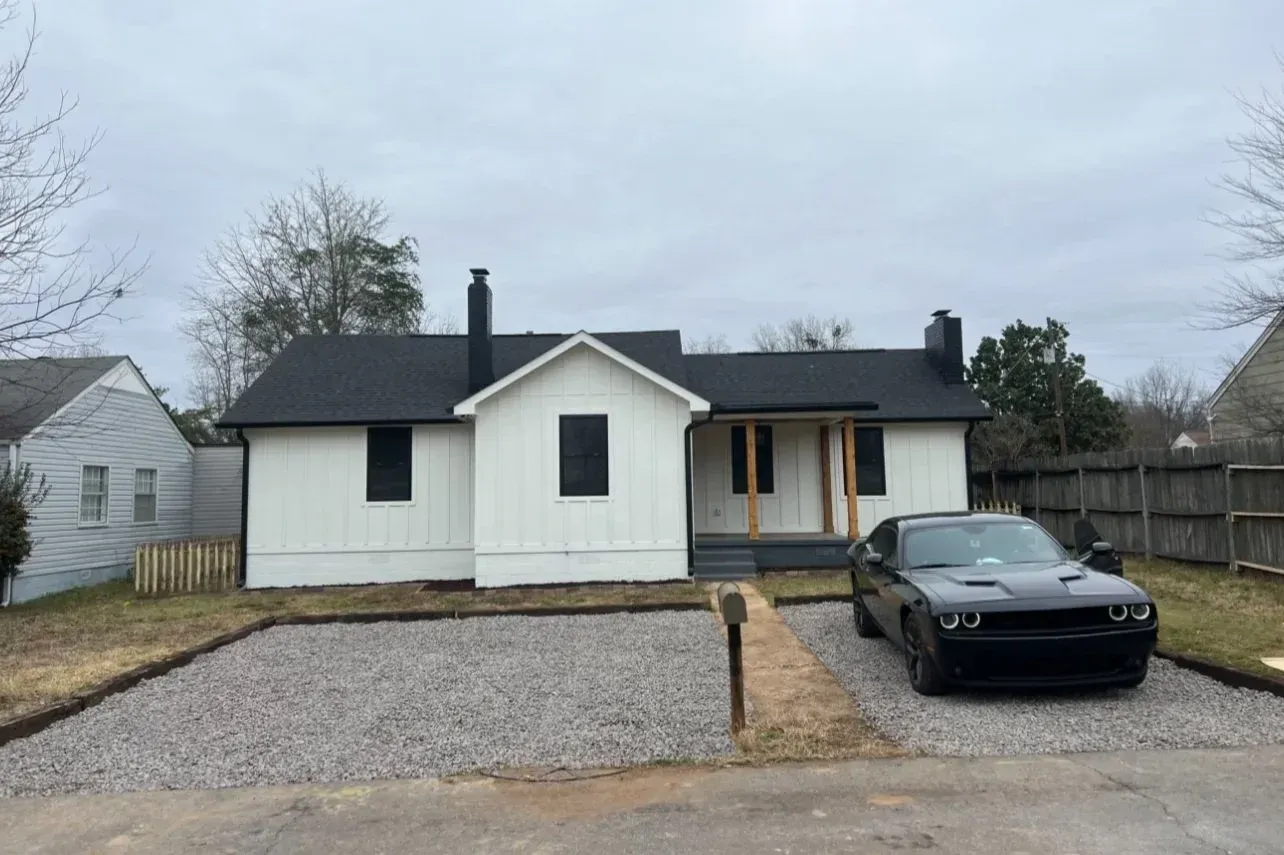 White house with black roof and accents, black car in driveway, gravel yard.