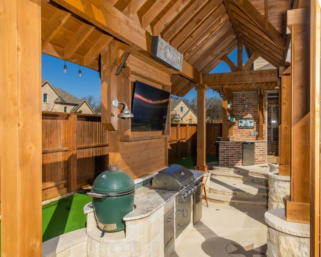 Outdoor kitchen with a green Big Green Egg grill, stainless steel appliances, stone counters, and a wall-mounted TV.