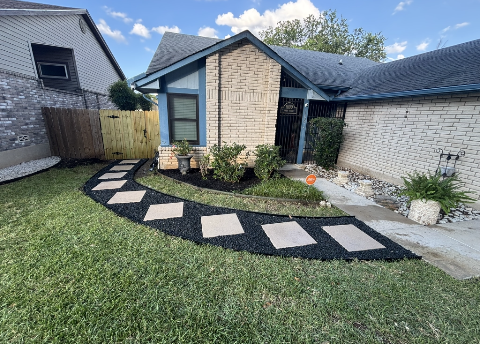 A curved stone walkway set in black mulch leads to the front entrance of a house with light stone and brick siding.