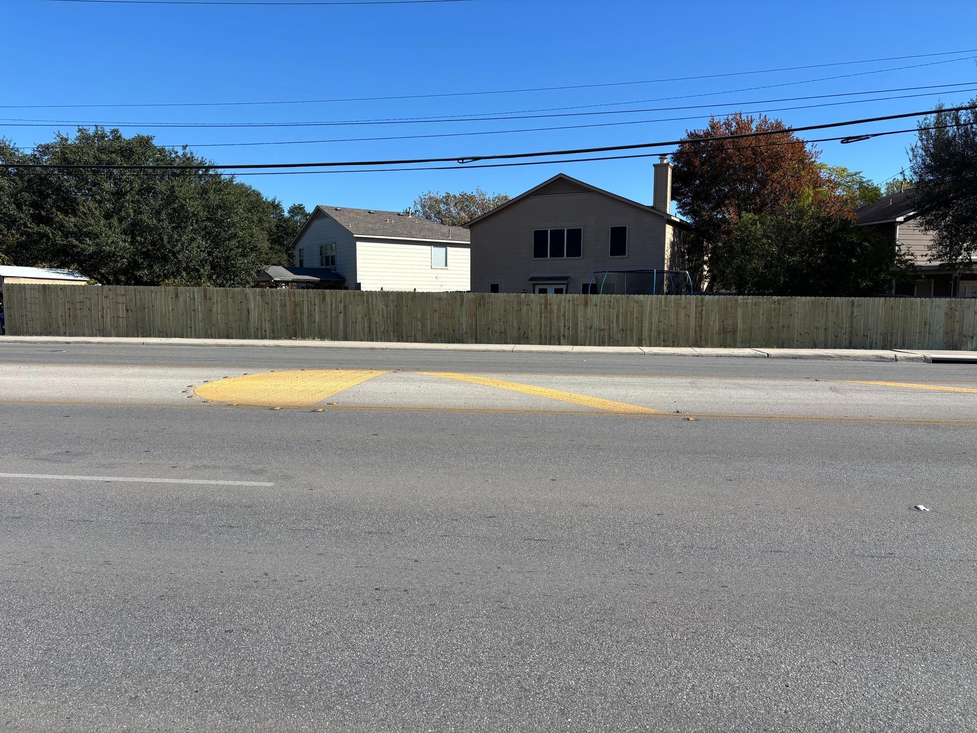 A wooden fence runs along the edge of a paved road with houses visible in the background under a clear blue sky.