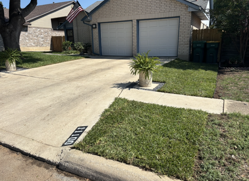 A house with a beige brick exterior, a two-car garage, and a concrete driveway with an address painted on the curb.