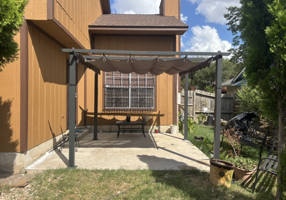 A dark grey metal pergola with a retractable fabric shade stands over a concrete patio next to a brown wooden house.