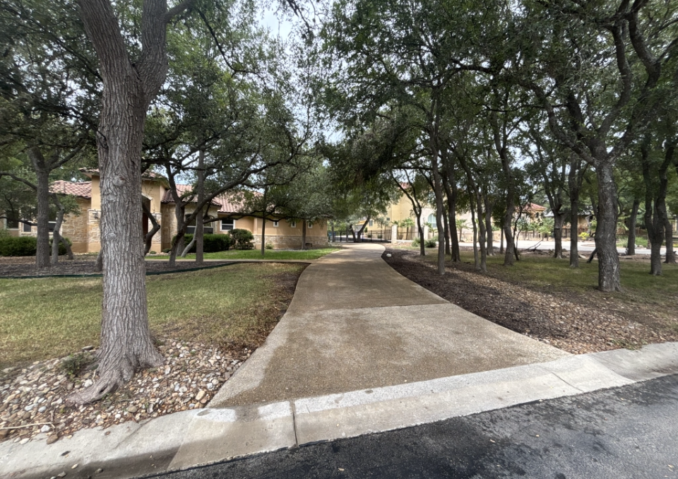 A paved driveway leads to a single-story stone house shaded by numerous mature trees in a residential neighborhood.