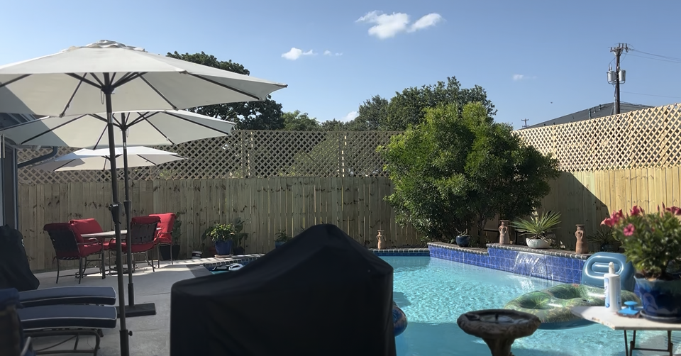 A backyard patio with white umbrellas, lounge chairs, a covered grill, and a blue swimming pool under a sunny sky.