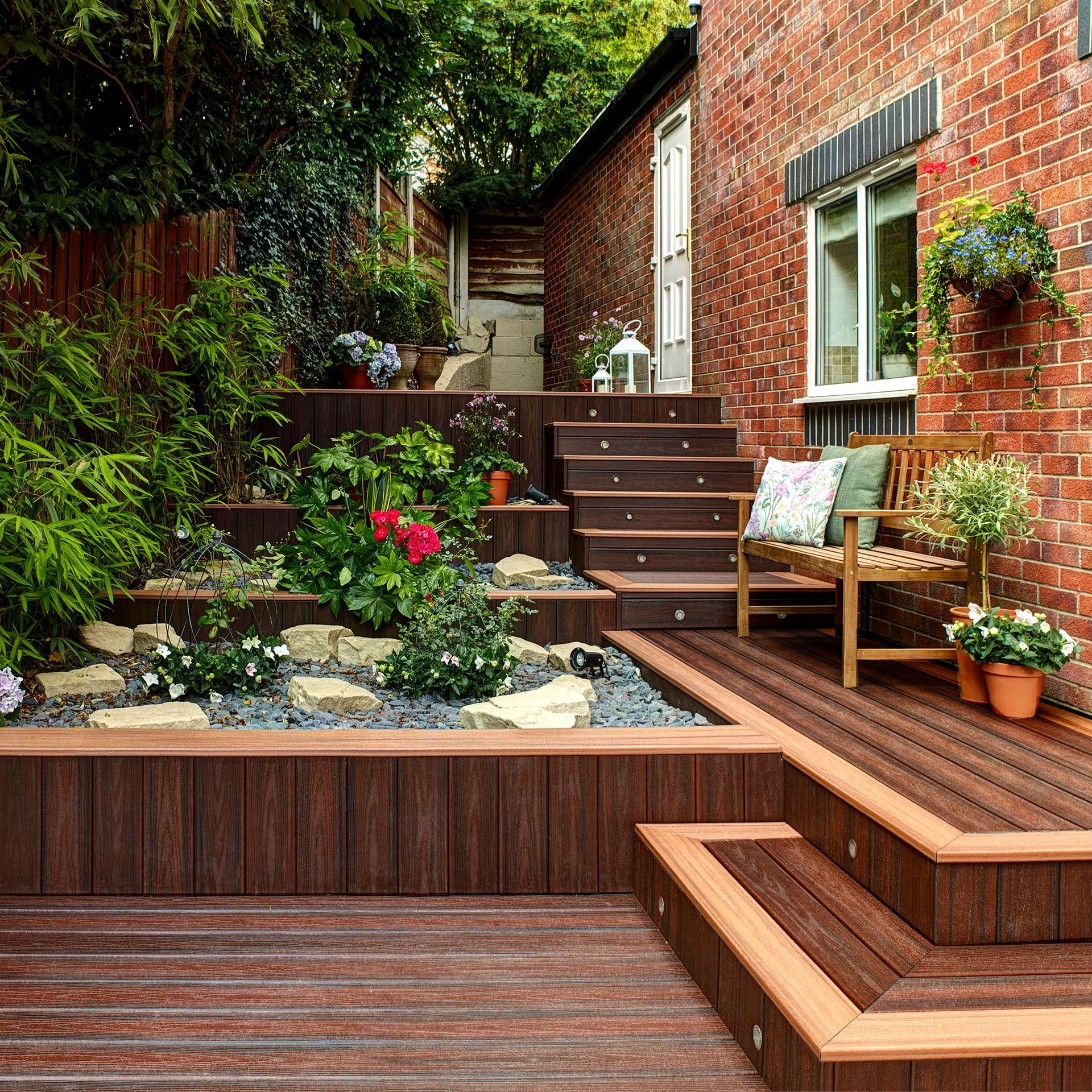 A multi-level outdoor wooden deck and stairs next to a brick house with flowerbeds and a wooden chair.