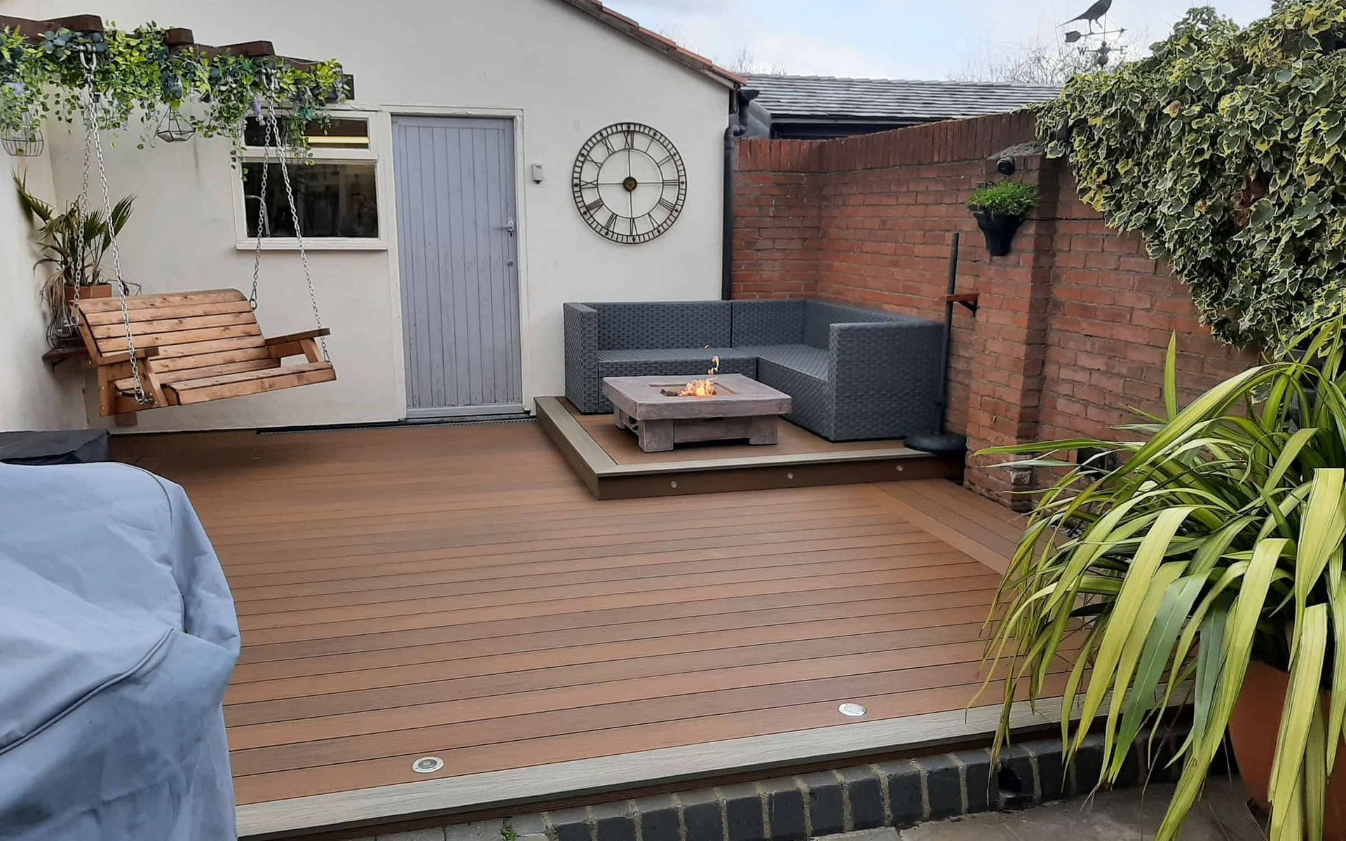 A backyard patio featuring a wood deck, a corner sofa with a fire pit, a hanging bench, and a brick wall.