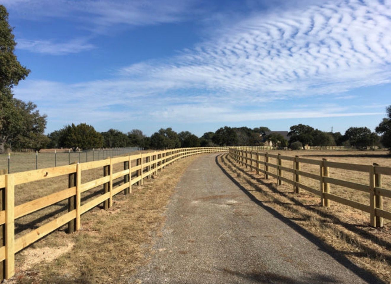 A paved driveway winds through a rural field, lined on both sides by wooden post-and-rail fences under a blue sky.
