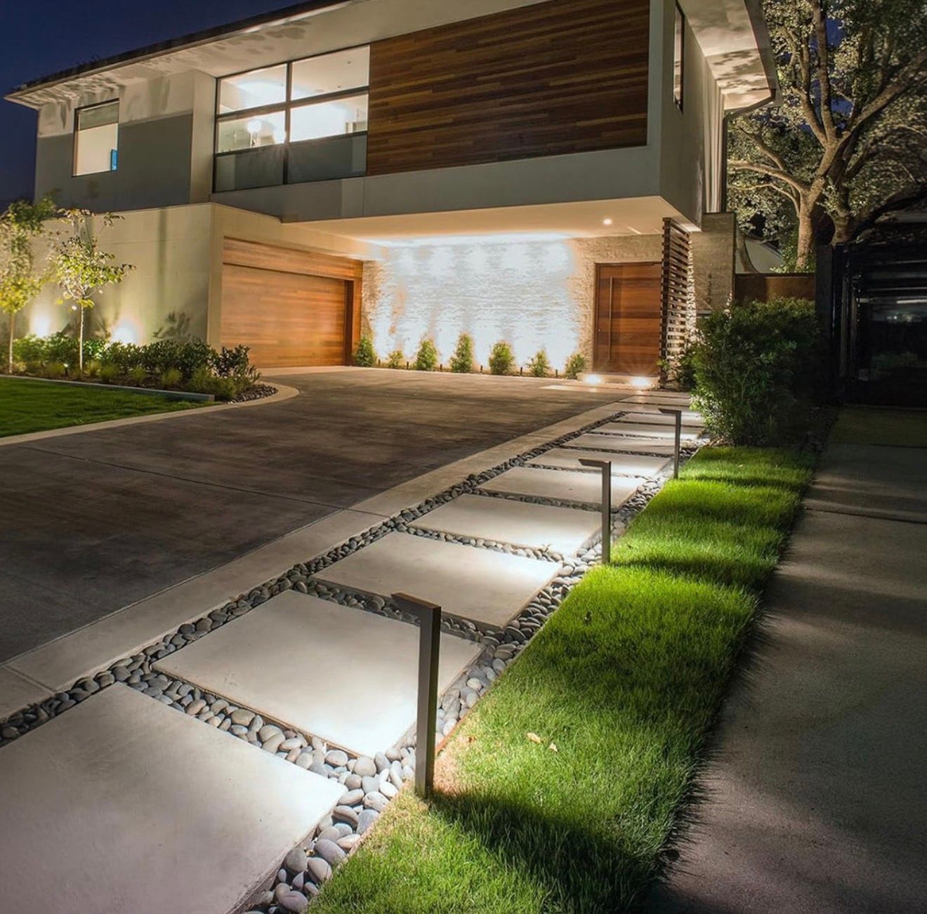 A modern home exterior at night with a stone pathway lit by path lights, leading to a wooden garage and front door.