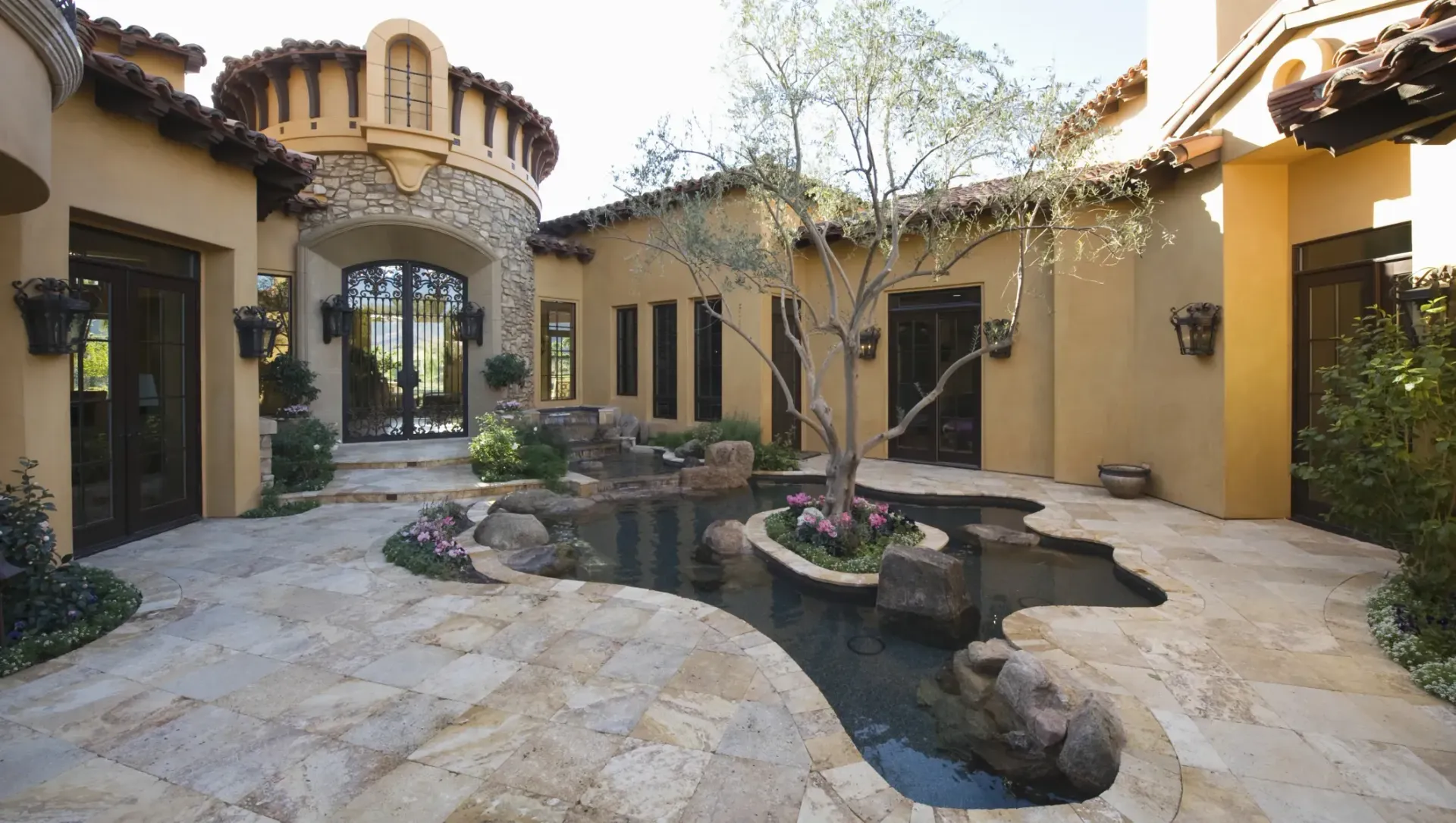 A Spanish-style courtyard with a stone-lined pond, a small tree, and warm-toned stucco buildings under a bright sky.