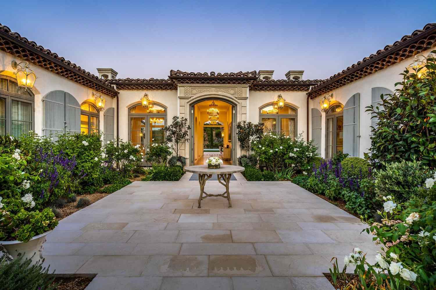 A stone courtyard with a central table leads to the arched entryway of a light-colored Mediterranean villa at dusk.