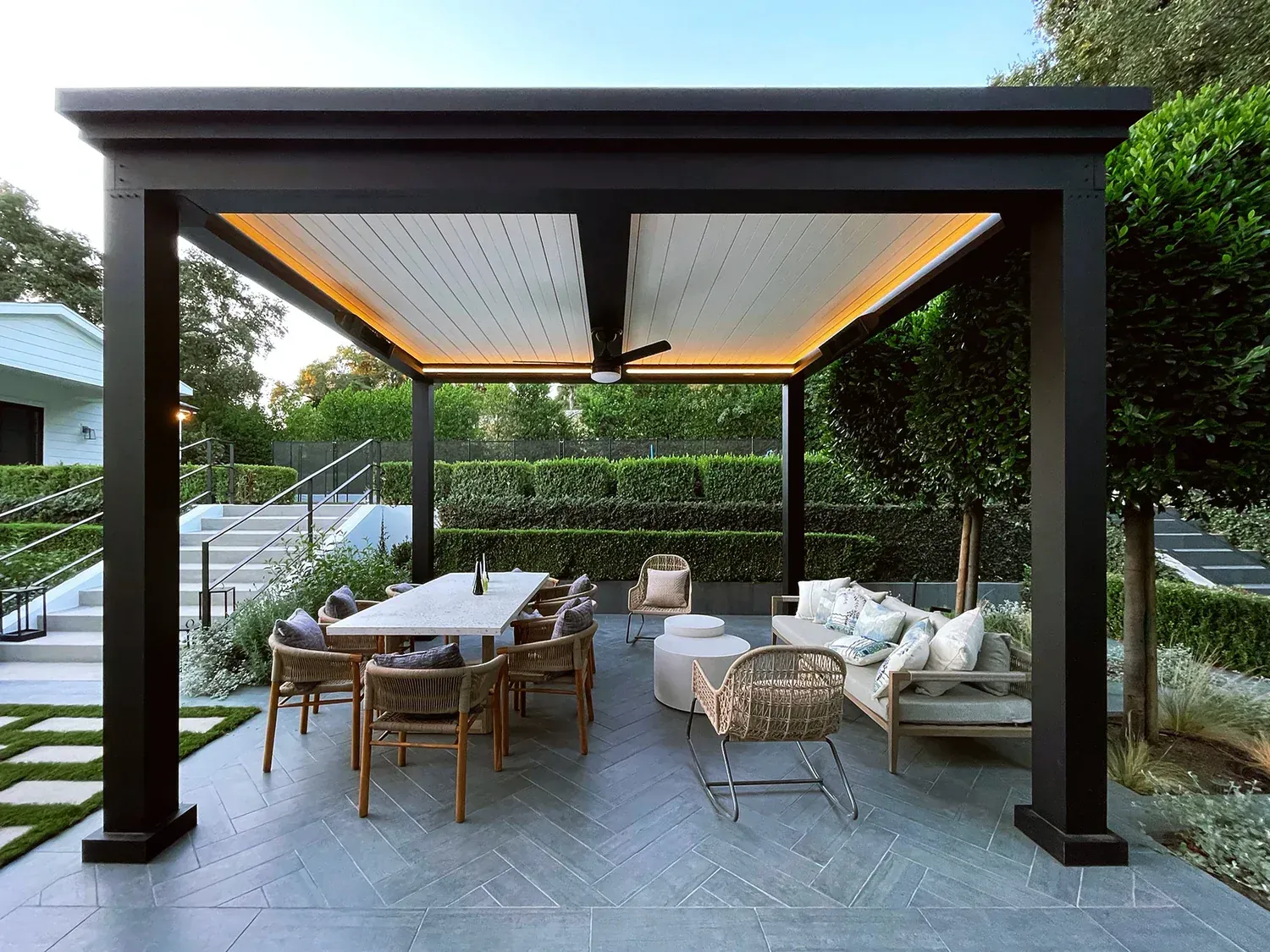 A modern black outdoor pergola featuring a dining area and seating lounge on a stone patio in a lush backyard.