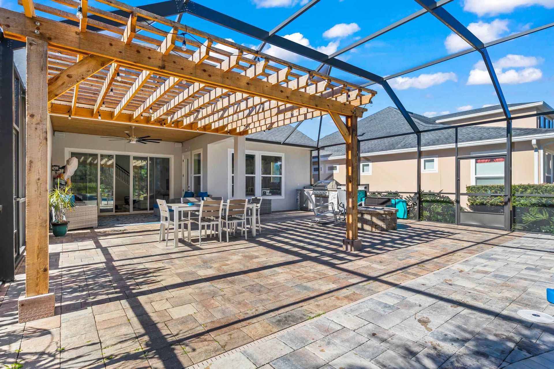 A paved patio with a wooden pergola, outdoor dining set, and screened-in enclosure under a bright, sunny sky.