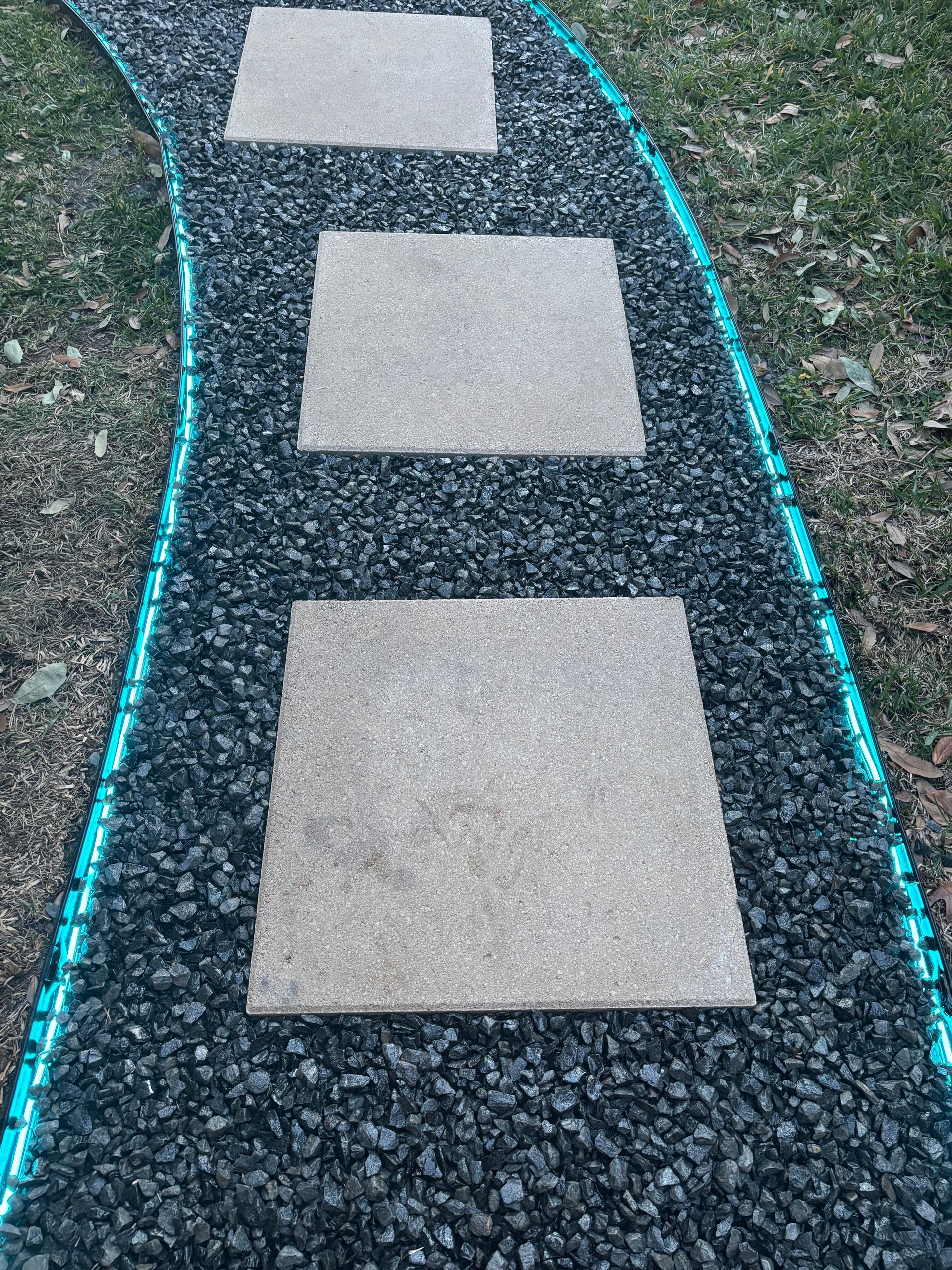 A curved garden path of dark gray gravel with square stone stepping stones, bordered by glowing teal LED light strips.