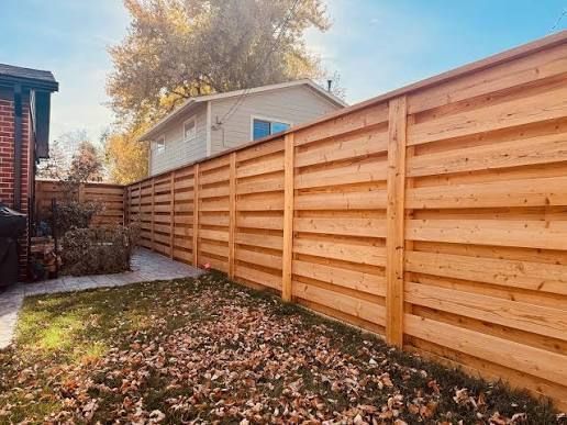 A horizontal wooden fence runs along the edge of a backyard covered in fallen leaves under a blue, autumn sky.