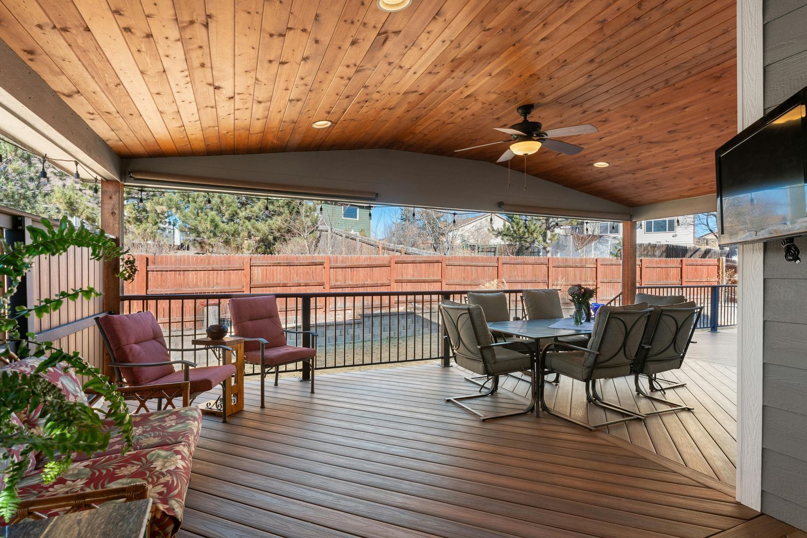 Covered wooden deck with seating, dining table, and a television. Backyard view.