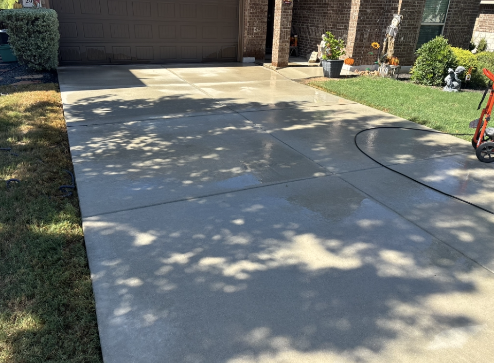 A clean, wet concrete driveway in front of a brick house, partially shaded by tree leaves, with a lawn on the side.