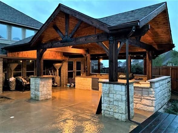 An outdoor patio with a dark wood-framed roof covering a built-in stone kitchen counter and bar area at twilight.