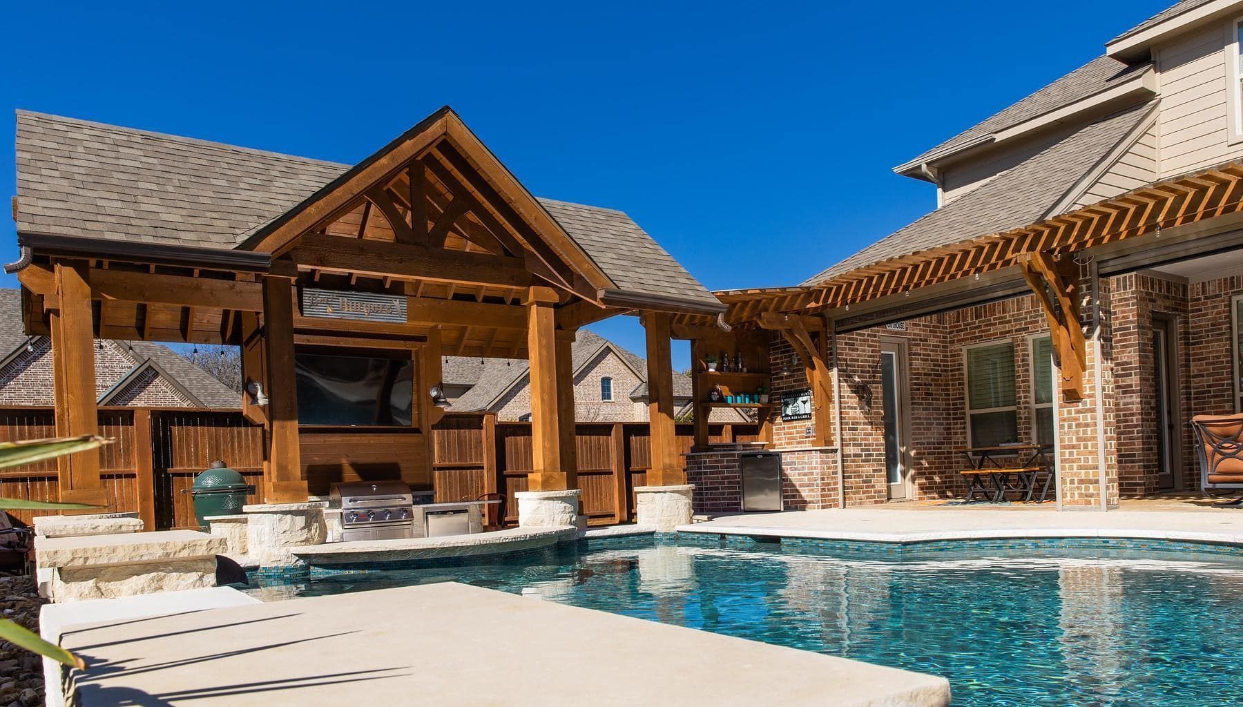 A backyard swimming pool with a stone patio, featuring a wooden pavilion with an outdoor kitchen next to the house.