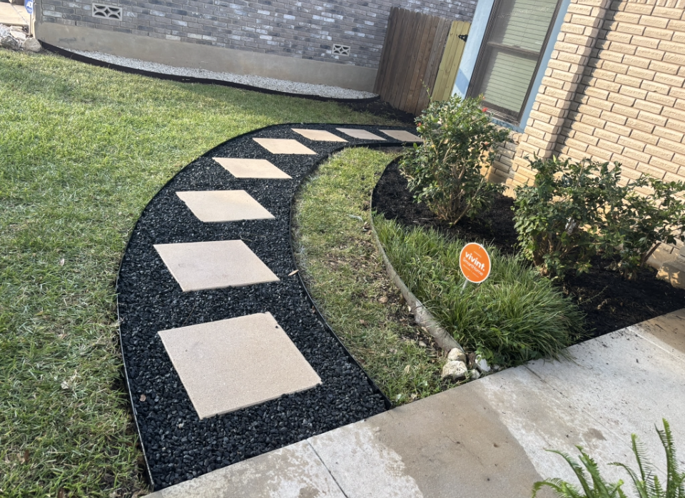 Curved garden path with square stone pavers set in black mulch next to a grassy lawn and brick wall.