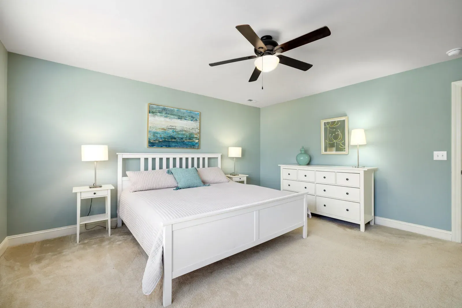 A bedroom with light blue walls, white furniture, a bed with white bedding, and a ceiling fan over a beige carpet.