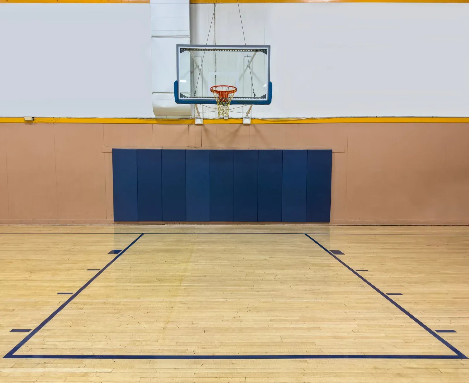 Basketball hoop mounted on a wall above blue padded panels and a wooden court with marked lines.
