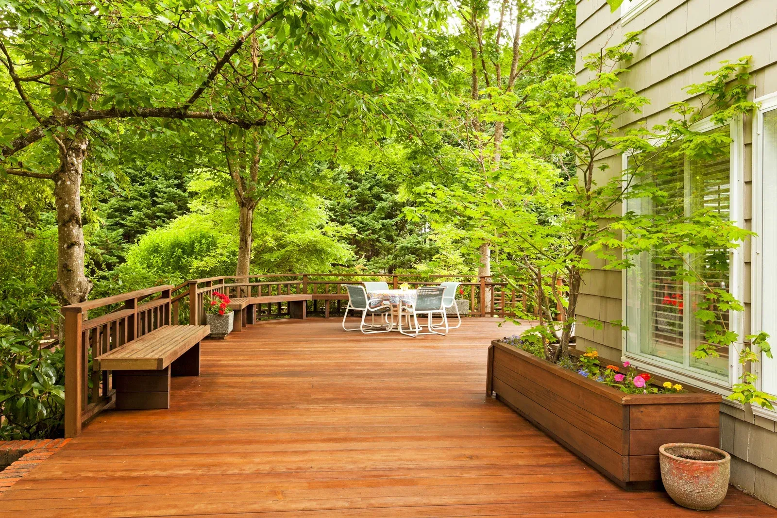 Wooden deck with benches, table, and chairs surrounded by trees, next to a house.