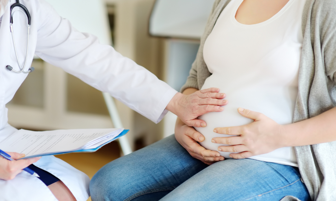 Pregnant woman in light blue dress holding yellow flowers in a sunny park.