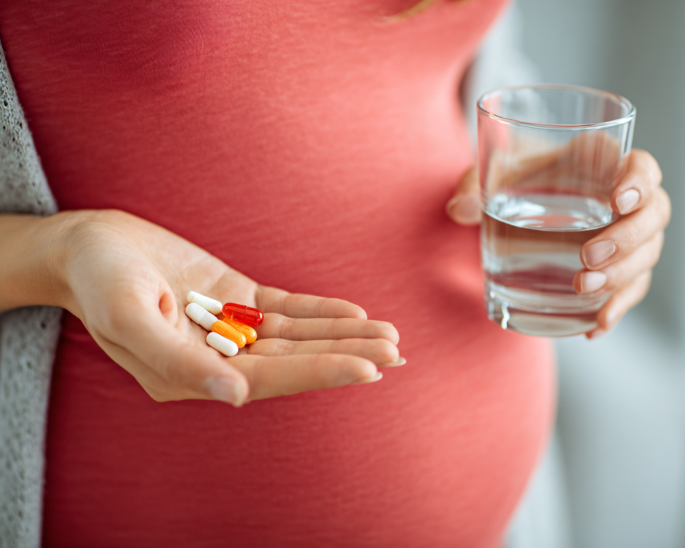 Pregnant woman holding pills in one hand and a glass of water in the other, indoors.
