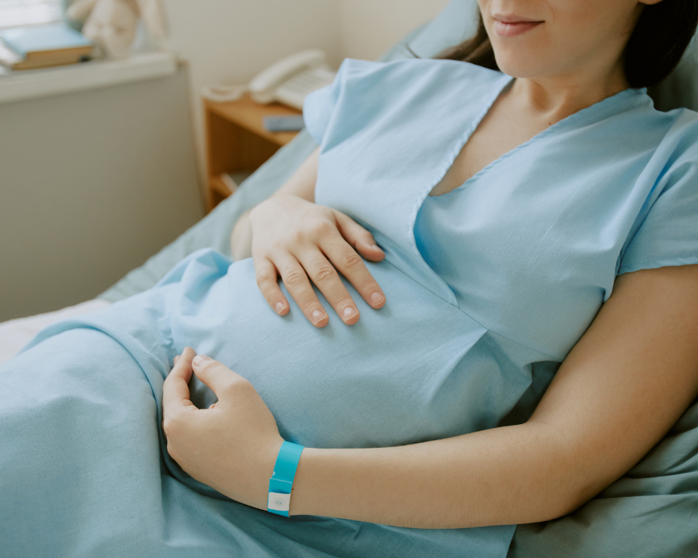 Pregnant woman in a hospital bed, wearing a blue gown, holding her stomach, with a hospital wristband.