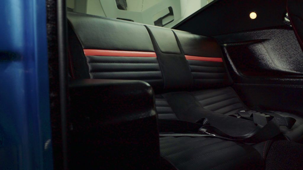 Interior of a classic car: black seats with red accents, visible seatbelts, and blue door panel.