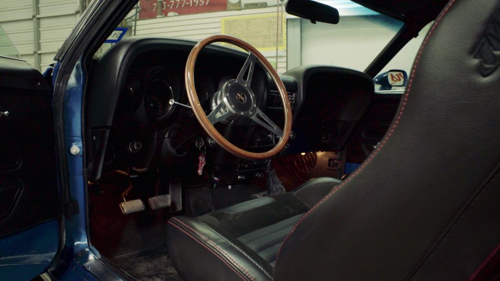 Interior of a classic blue car with a wooden steering wheel, black seats, and red stitching.