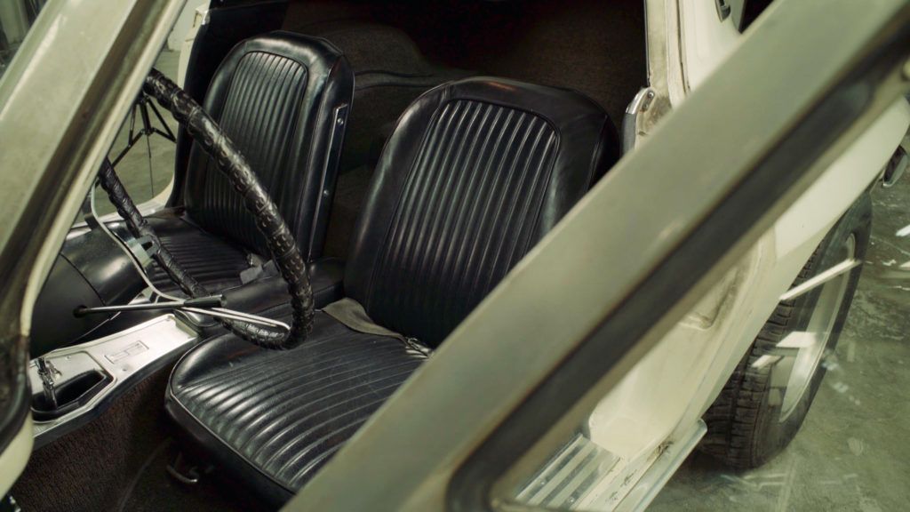 Interior of a vintage white car: black seats, steering wheel, and open door.
