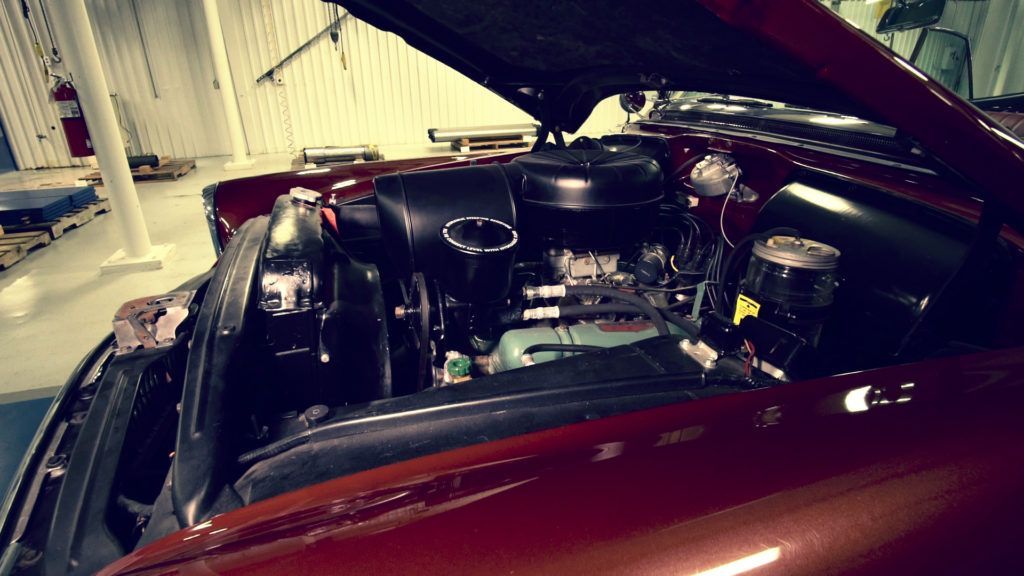Red classic car engine bay, open hood, in a garage.