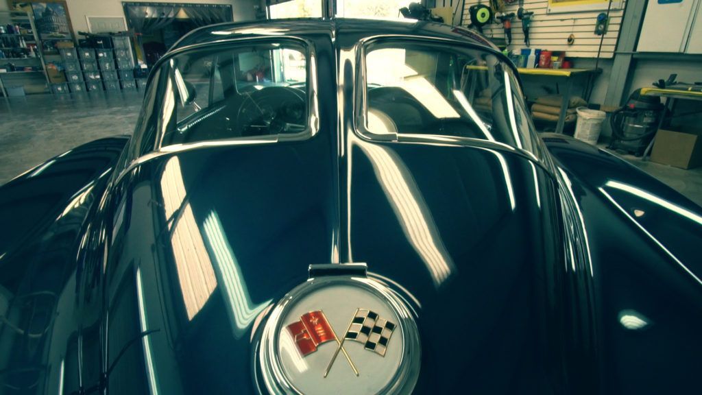 Dark blue vintage car, hood ornament with Corvette emblem, inside a workshop.