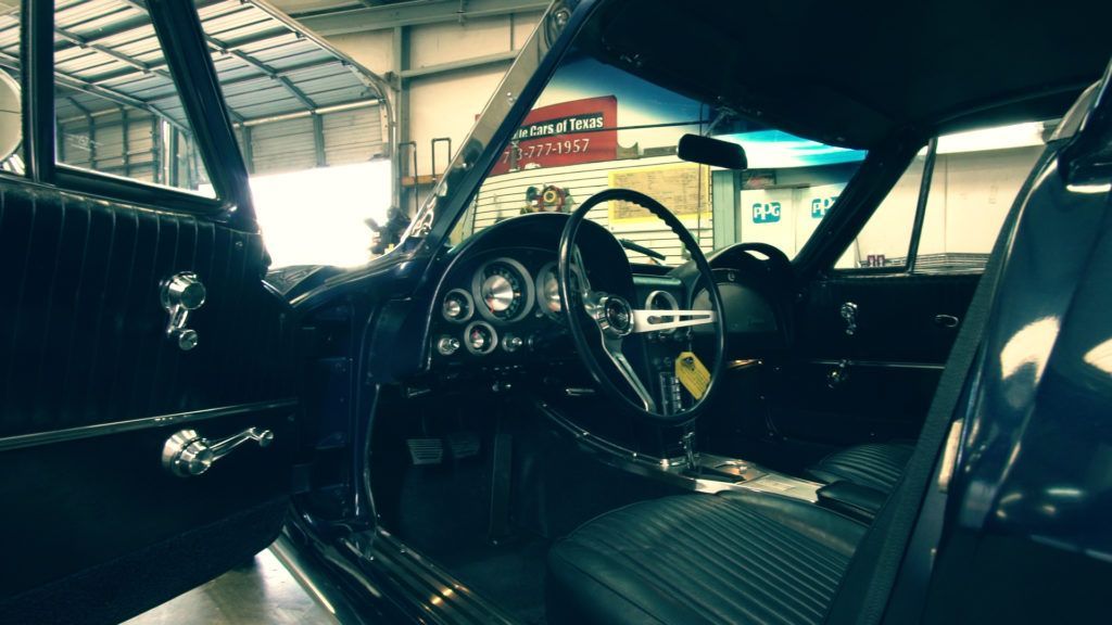Interior of a classic black car. Driver's side door open, black steering wheel, and dashboard visible.