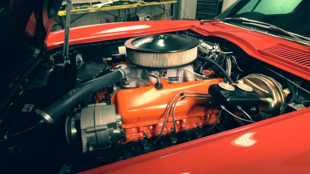 Red classic car engine bay, orange engine, black air filter and intake, parked indoors.