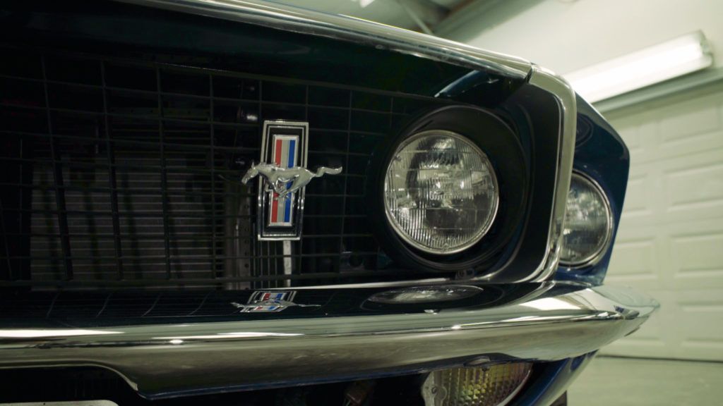 Close-up of a classic blue Ford Mustang front end, showing the grill, headlights, and emblem in a garage.