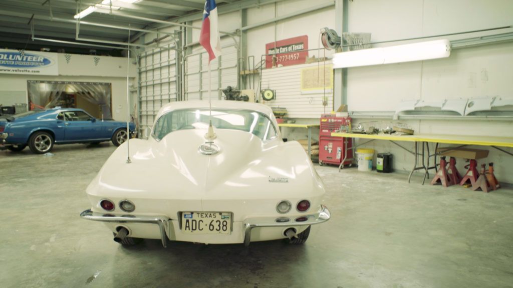 White vintage Corvette in a garage with a blue car in the background.