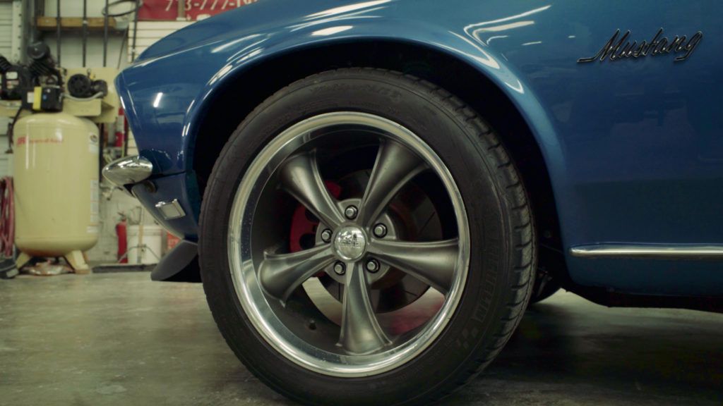 Blue classic Mustang's front wheel and fender in a garage, showing details of the rim and brakes.