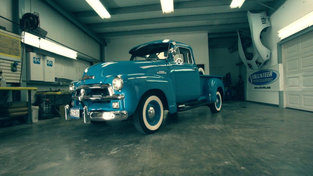 Blue vintage pickup truck inside a garage, with white tires and a light shining on it.