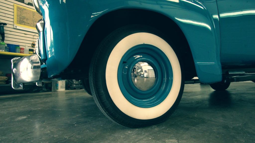 Blue classic car, white-walled tire, chrome hubcap, parked on a gray concrete floor, in a shop.