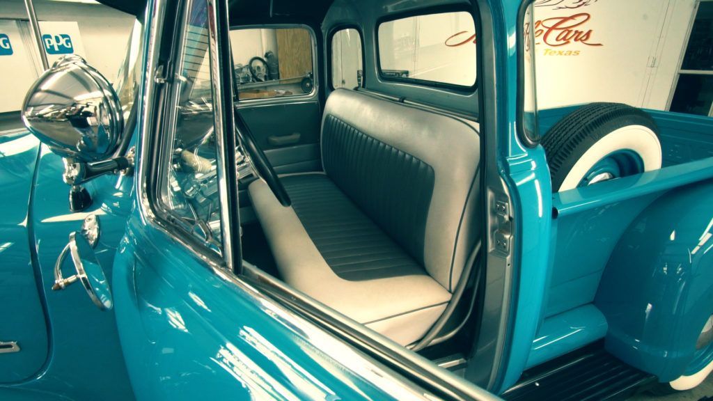 Teal classic pickup truck interior; light and dark gray bench seat, chrome accents, white-wall spare tire.